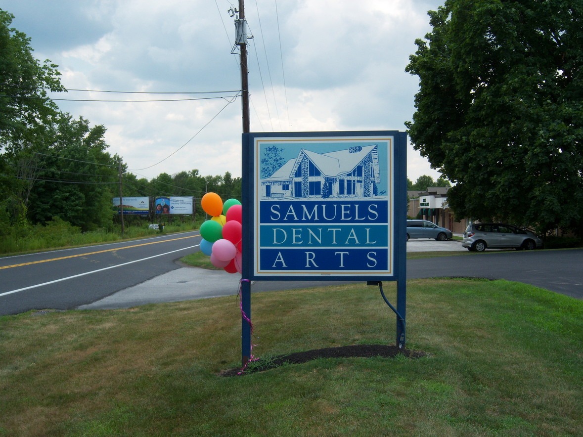 A sign for Samuels Dental Arts stands on a grassy area beside a road, with colorful balloons attached to it.