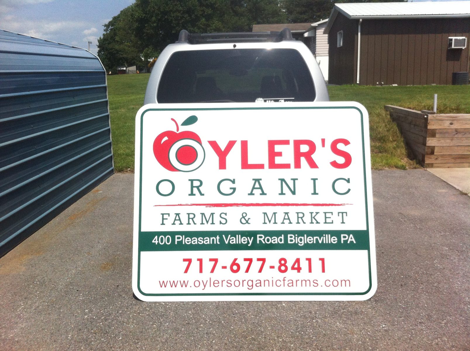A sign for Oyler's Organic Farms & Market stands in front of a parked vehicle in an outdoor area.