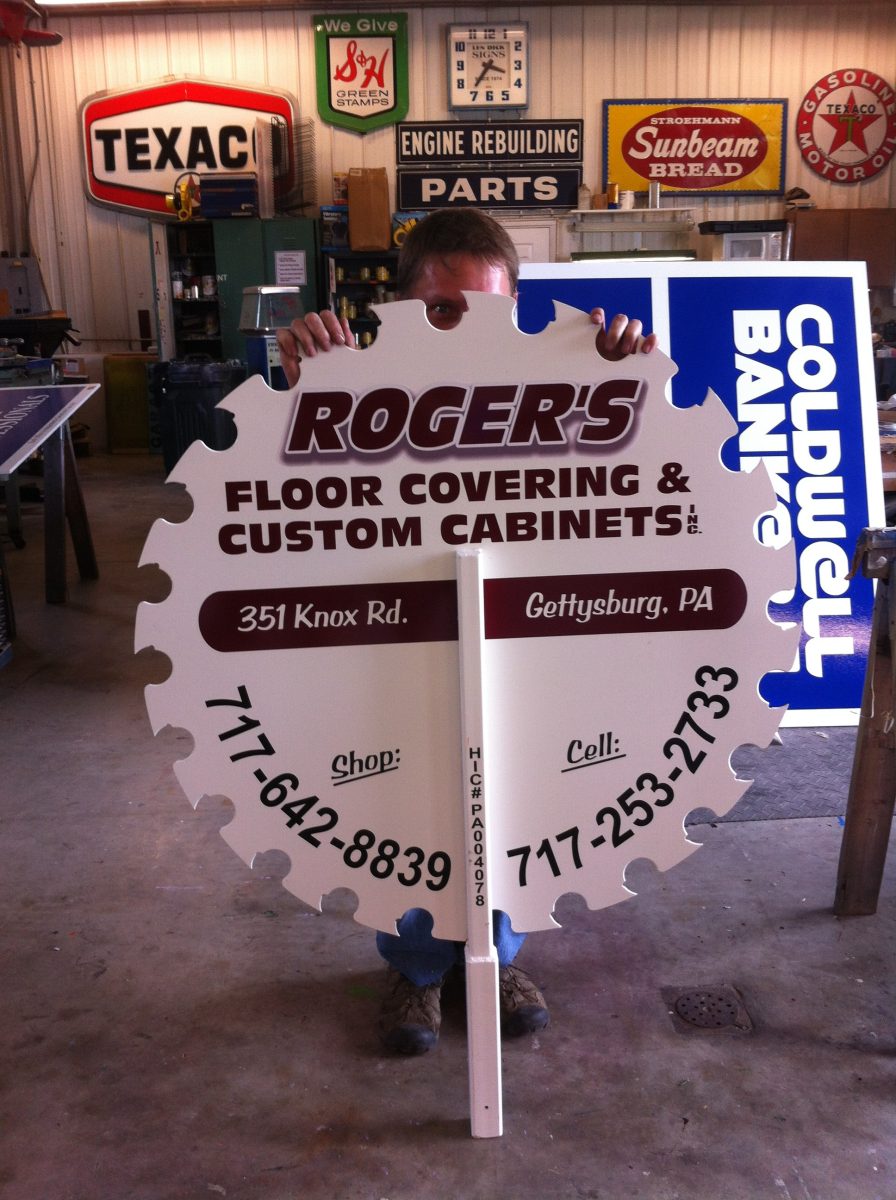 Person holding a large saw blade-shaped sign for Roger's Floor Covering & Custom Cabinets in a workshop with vintage signs.