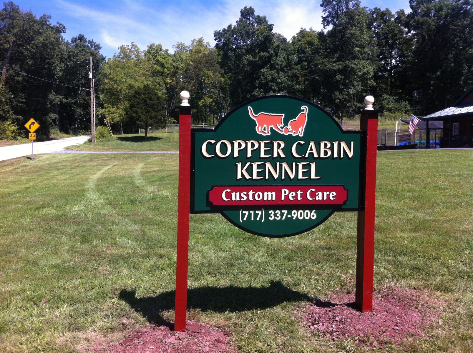 A sign for Copper Cabin Kennel offering custom pet care, with a phone number, stands on a grassy lawn near a road.