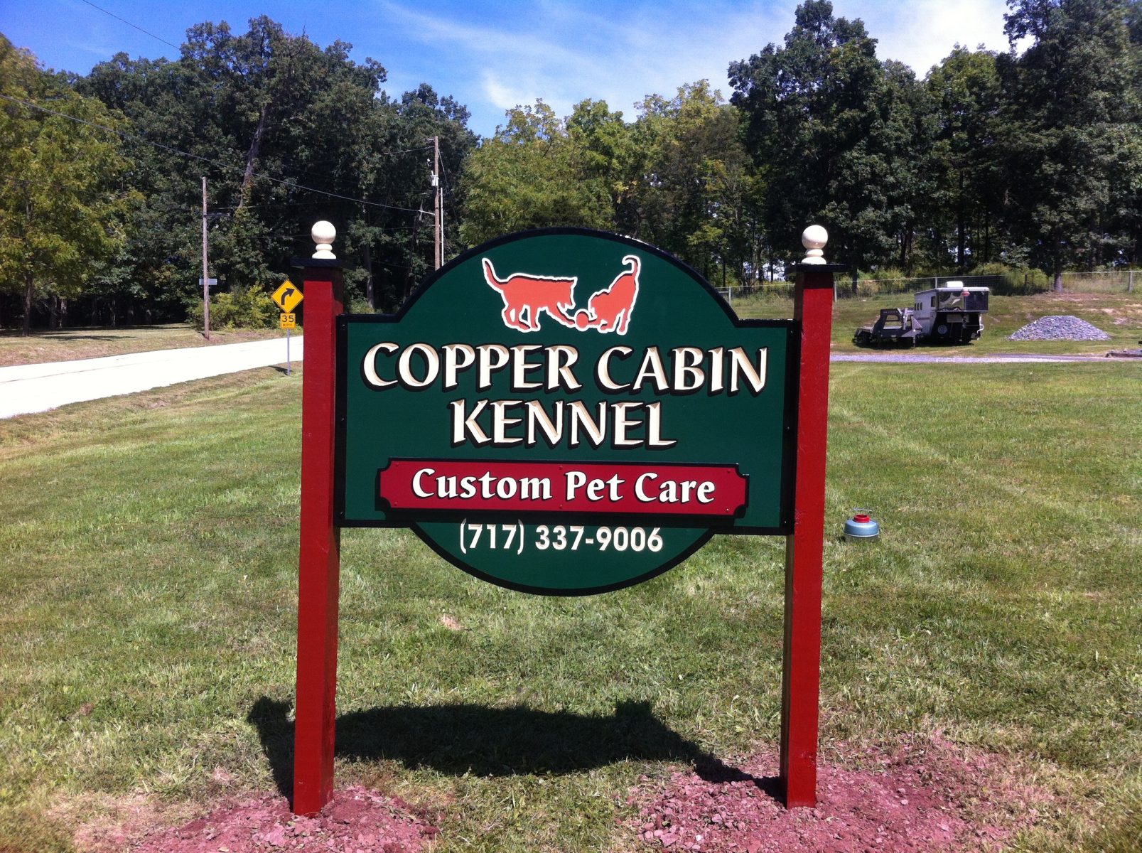 A green and red sign reads "Copper Cabin Kennel, Custom Pet Care, (717) 337-9006" on a grassy area near a road.