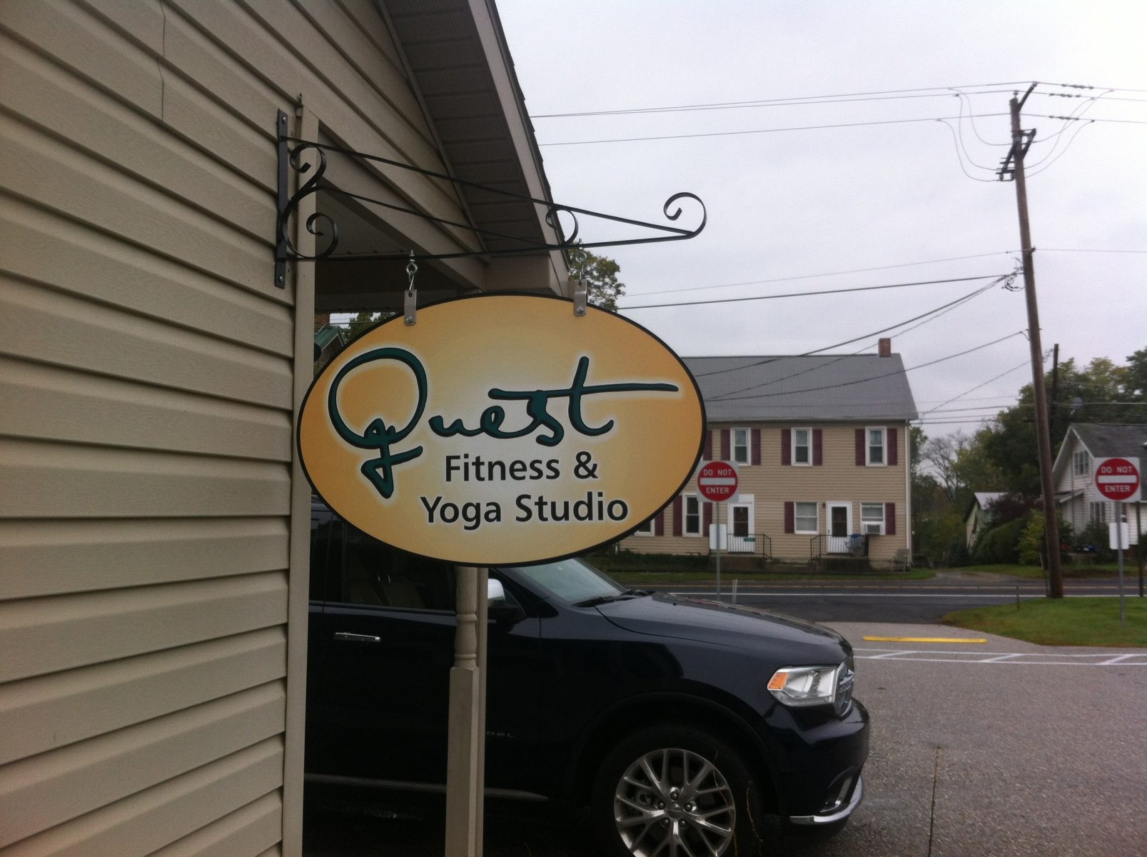 Oval sign reading "Quest Fitness & Yoga Studio" hangs on the exterior of a building near a street and parked car.