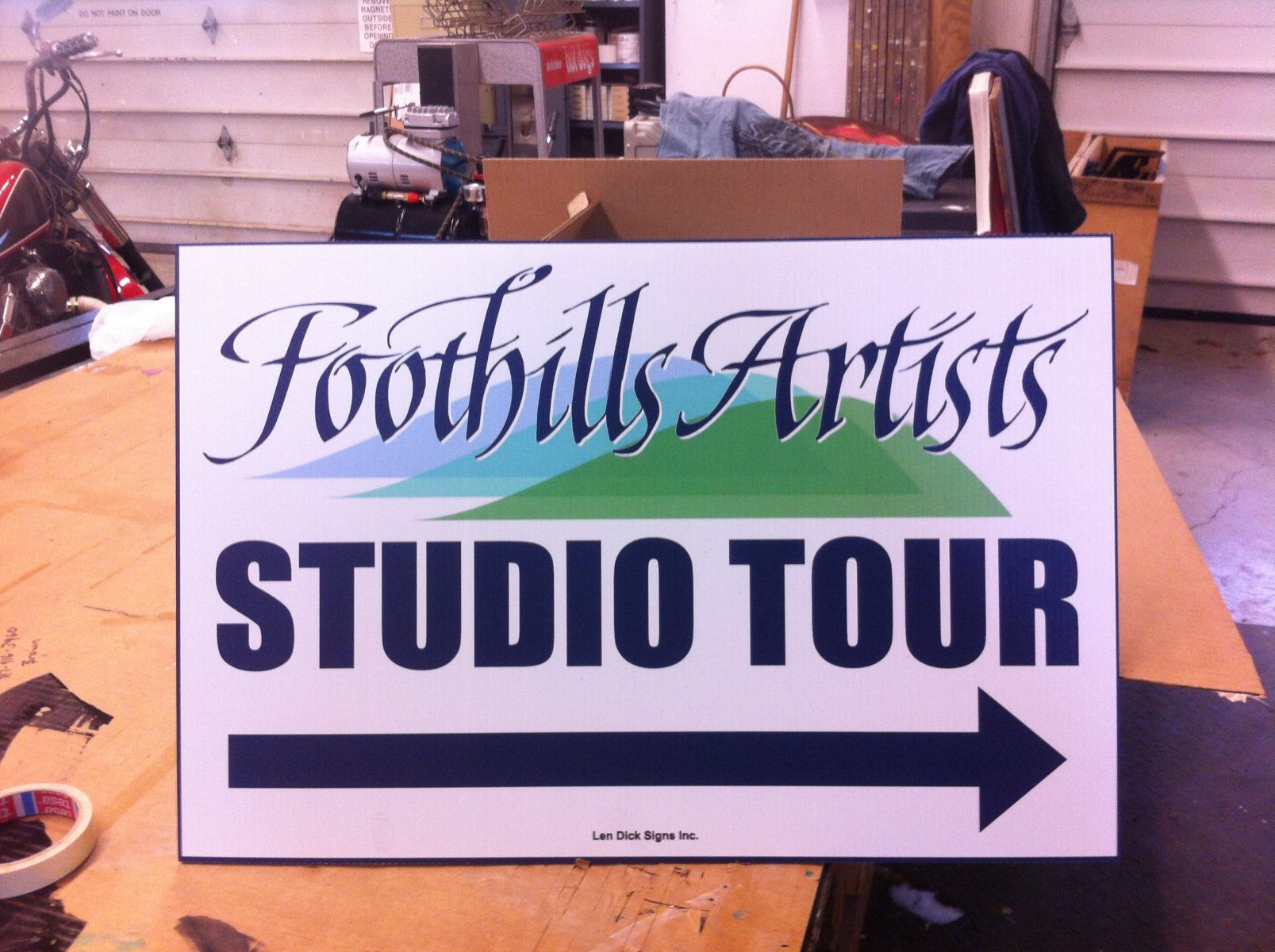 A sign reads "Foothills Artists Studio Tour" with a right-pointing arrow, placed on a workbench in a workshop.