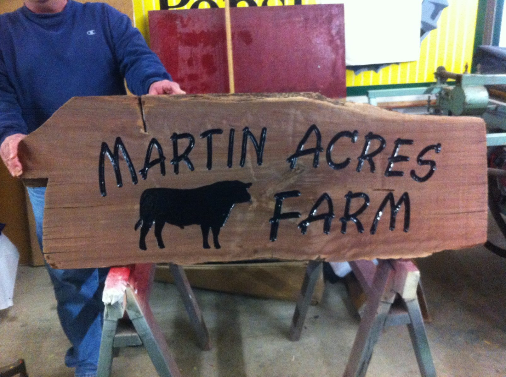 Wooden sign reads "Martin Acres Farm" with a silhouette of a cow, held by a person indoors.