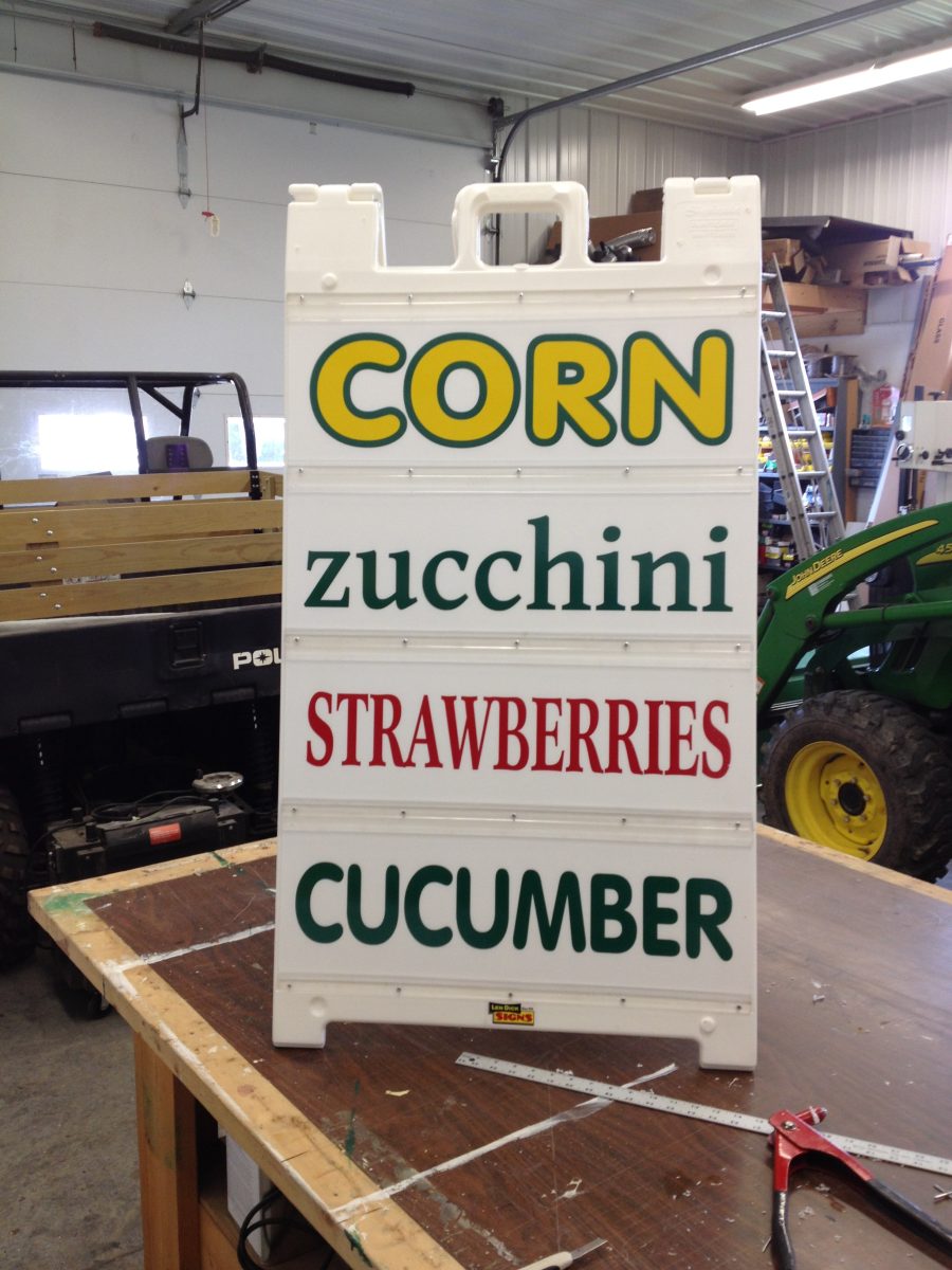 A white sandwich board sign lists corn, zucchini, strawberries, and cucumber for sale in a garage setting.