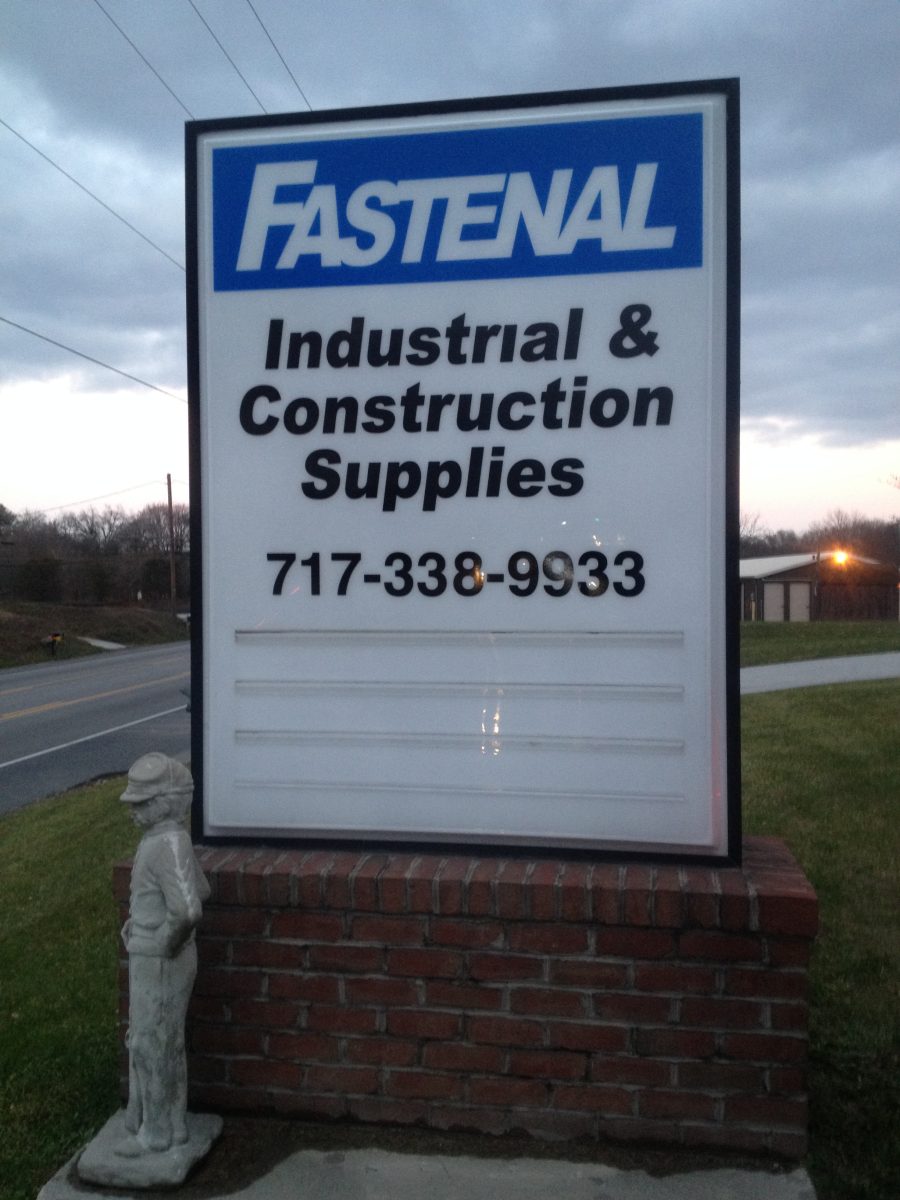 Sign for Fastenal Industrial & Construction Supplies with phone number, mounted on brick, small statue in front.