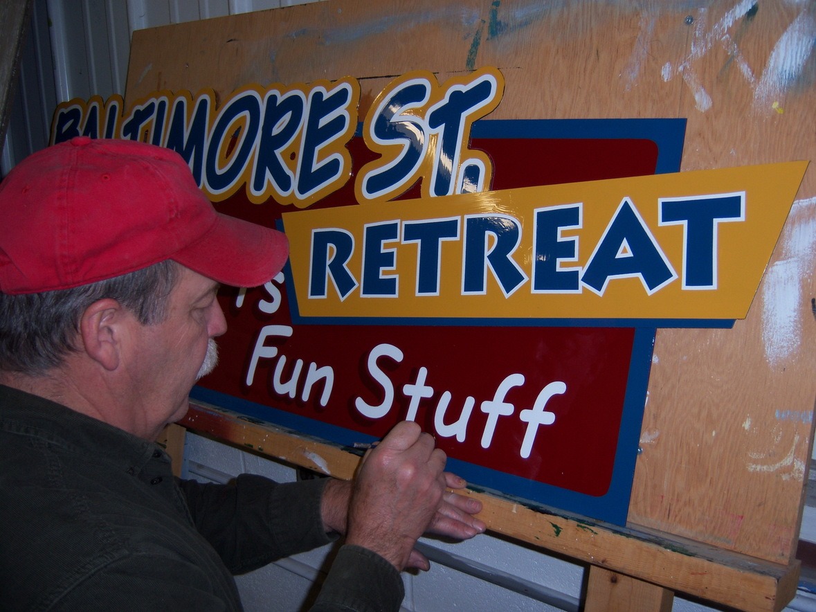 A person in a red cap paints letters on a colorful sign that reads "BALTIMORE ST. RETREAT Fun Stuff.