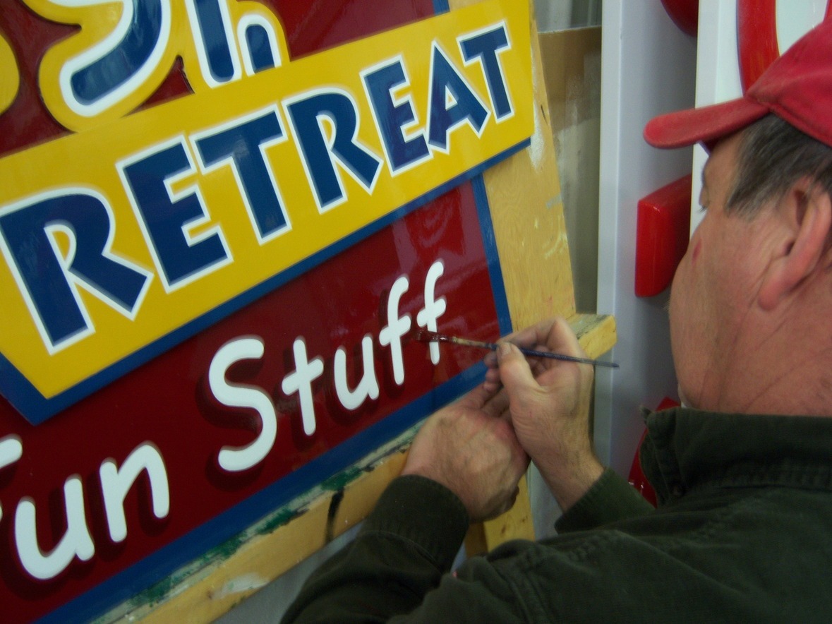 A person hand-paints letters on a colorful sign that reads "RETREAT Fun Stuff.