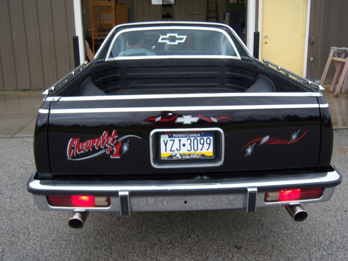 Rear view of a black Chevrolet El Camino with Pennsylvania plates and custom decals, parked outside a garage.